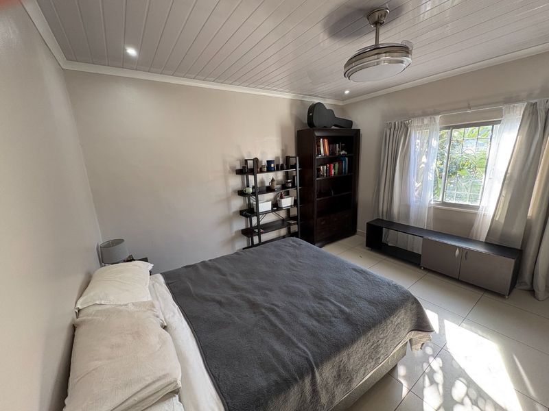 A bedroom featuring tiled flooring, a ceiling fan, and a window with security bars, providing a functional and well-lit living space.