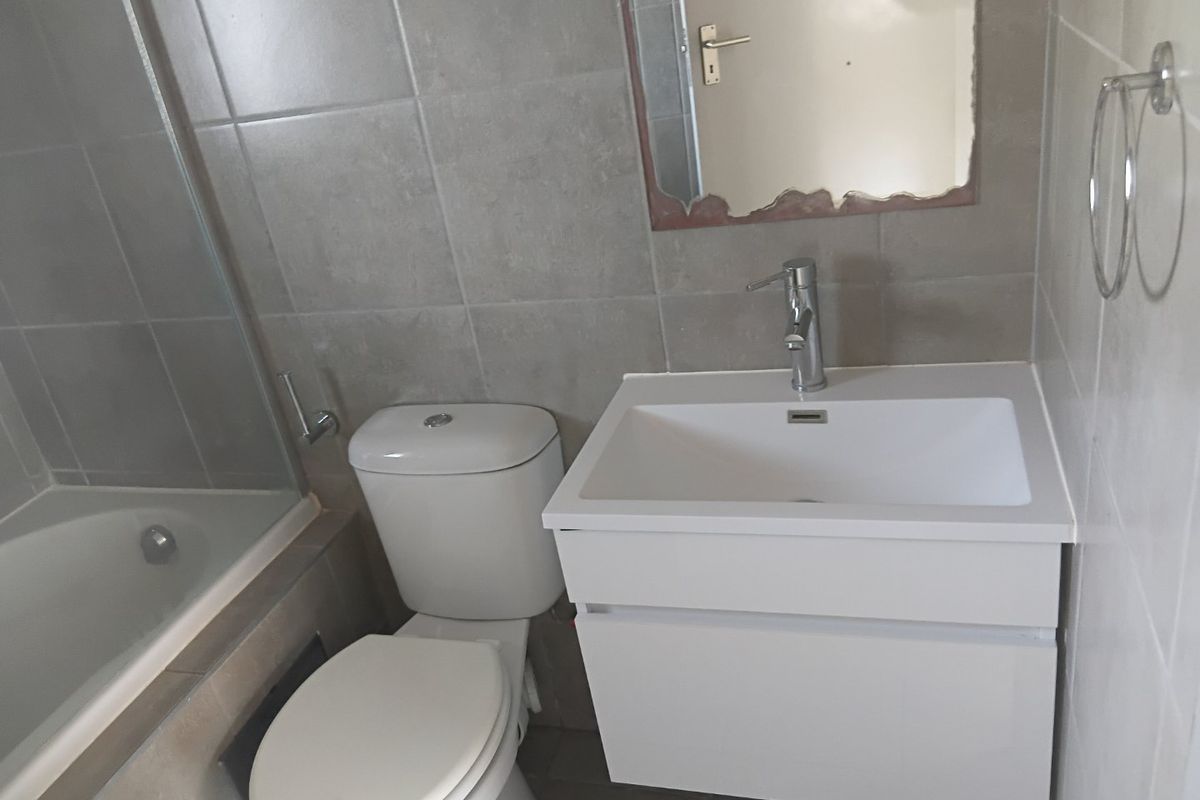 A compact bathroom featuring a modern white vanity, toilet, and a bathtub with a glass shower screen, finished with neutral grey wall tiles.