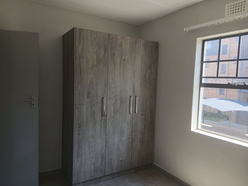 A clean, empty bedroom featuring a modern wood-finish built-in wardrobe, tiled flooring, and a window with security bars.
