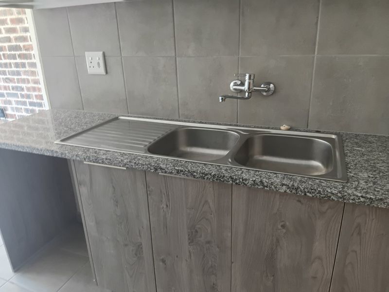 A clean, modern kitchen area featuring a stainless steel double sink, granite countertop, and wood-look cabinetry with a neutral tiled backsplash.