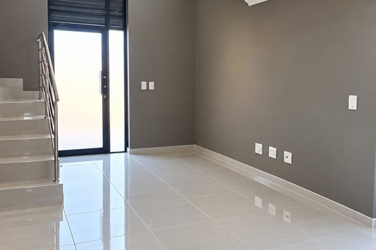 A bright, modern, and empty living area featuring polished tiled floors, recessed lighting, and a sleek staircase with a metal balustrade.