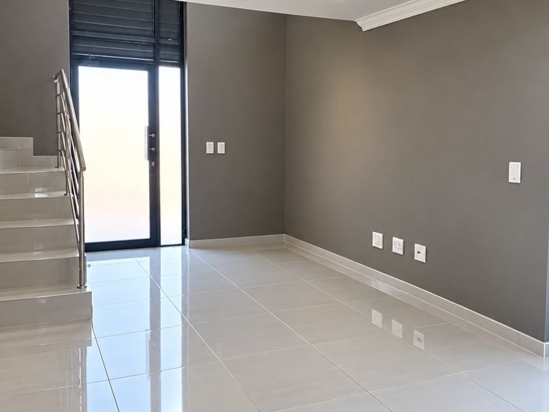 A bright, modern, and empty living area featuring polished tiled floors, recessed lighting, and a sleek staircase with a metal balustrade.