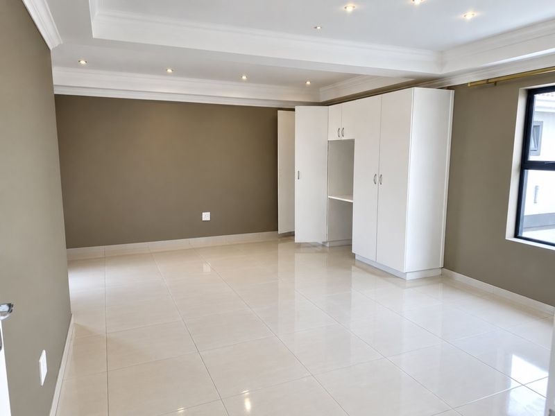 A spacious, empty bedroom featuring neutral wall tones, polished tiled flooring, recessed ceiling lights, and ample built-in cupboard storage.