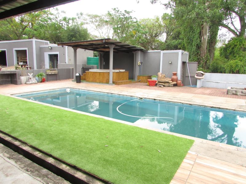 Outdoor pool area featuring a swimming pool, covered patio with a built-in jacuzzi, paved decking, and artificial lawn in a garden setting.