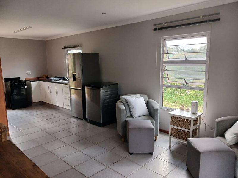 An open-plan living and kitchen area featuring tiled flooring, white cabinetry, and large louvered windows providing natural light and garden views.