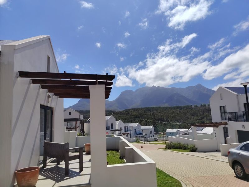 A modern residential exterior featuring a timber pergola, paved driveway, and white rendered walls, set against a scenic mountain backdrop.