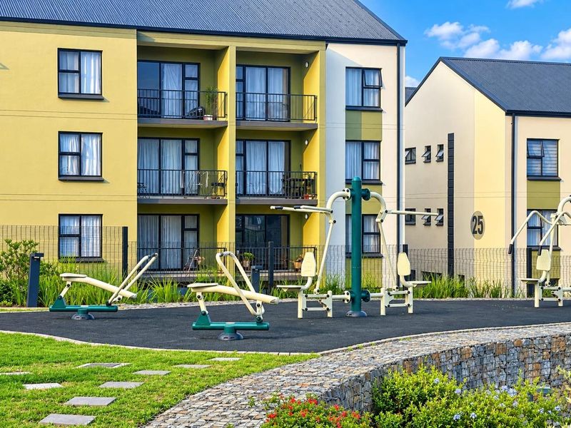 Outdoor fitness area featuring fixed exercise equipment on a rubberized surface, set within a well-maintained apartment complex with gabion wall landscaping.