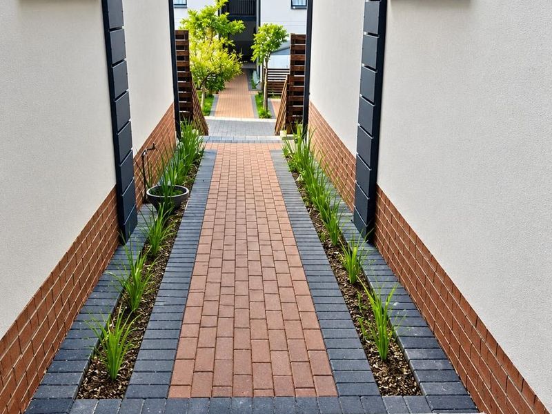 A well-maintained paved walkway between two apartment building sections, featuring neat brickwork, modern windows, and landscaped garden borders.