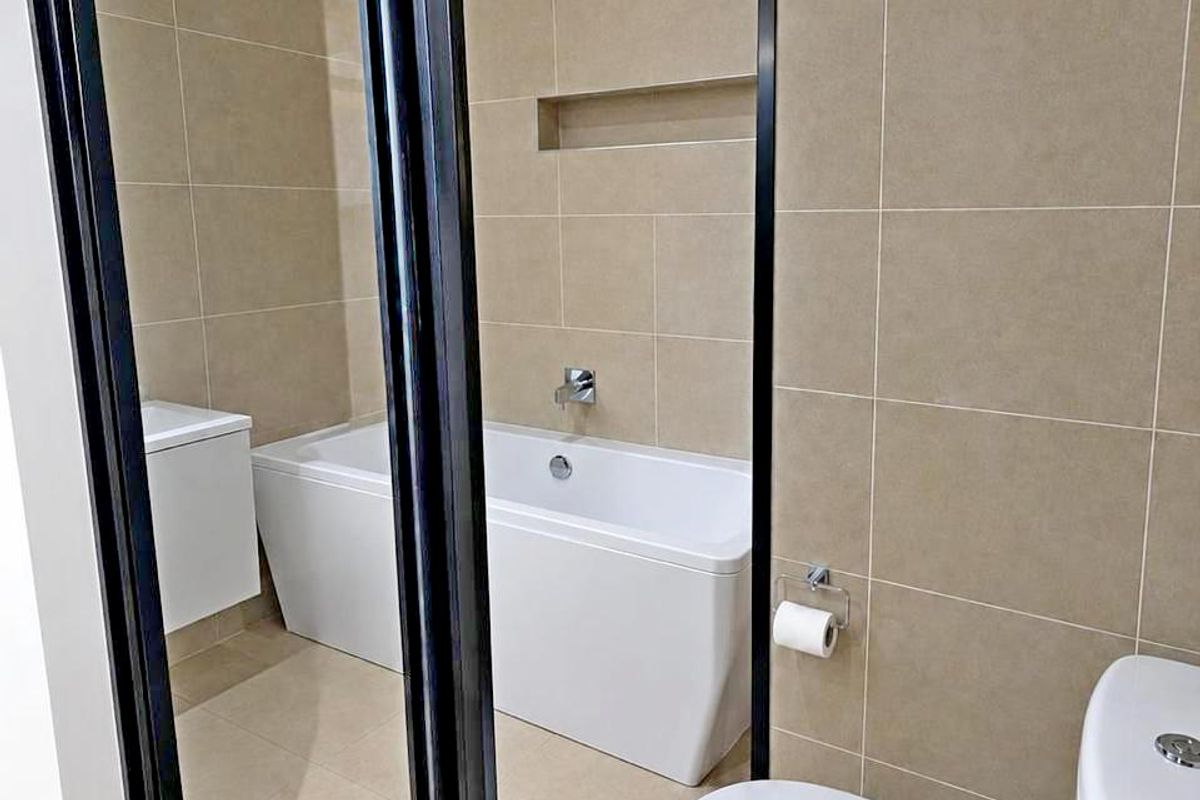 Modern bathroom featuring a sleek bathtub, wall-mounted vanity, and a contemporary toilet, finished with neutral wall and floor tiles and a black-framed glass partition.
