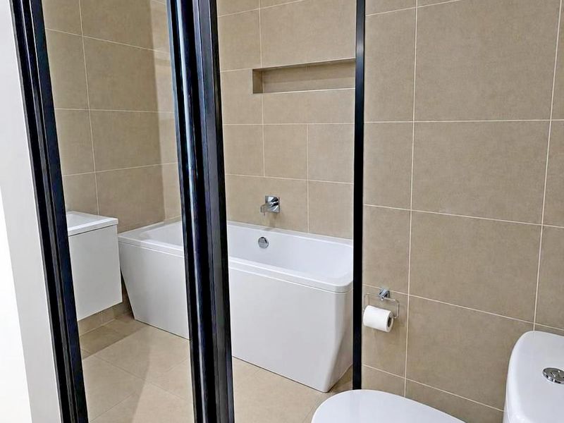 Modern bathroom featuring a sleek bathtub, wall-mounted vanity, and a contemporary toilet, finished with neutral wall and floor tiles and a black-framed glass partition.