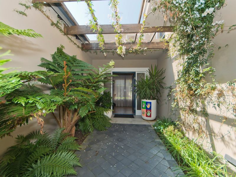 A charming covered entrance patio featuring a timber pergola with glass inserts, stone paving, and lush garden landscaping leading to the main entrance.