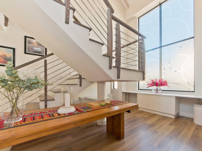 Bright entrance hall featuring a modern staircase with stainless steel balustrades, timber flooring, and built-in cabinetry under a large feature window.