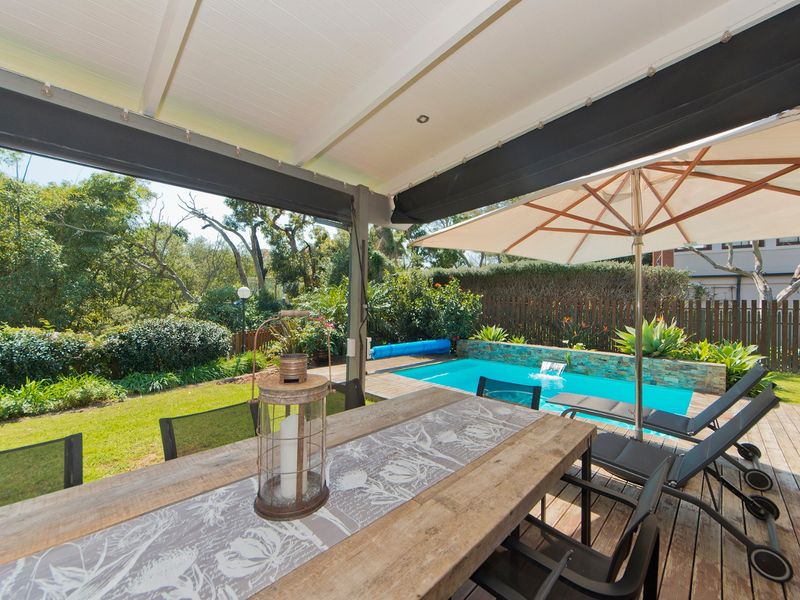 Covered patio area featuring timber decking and retractable blinds, overlooking a private swimming pool with a stone-clad water feature and landscaped garden.
