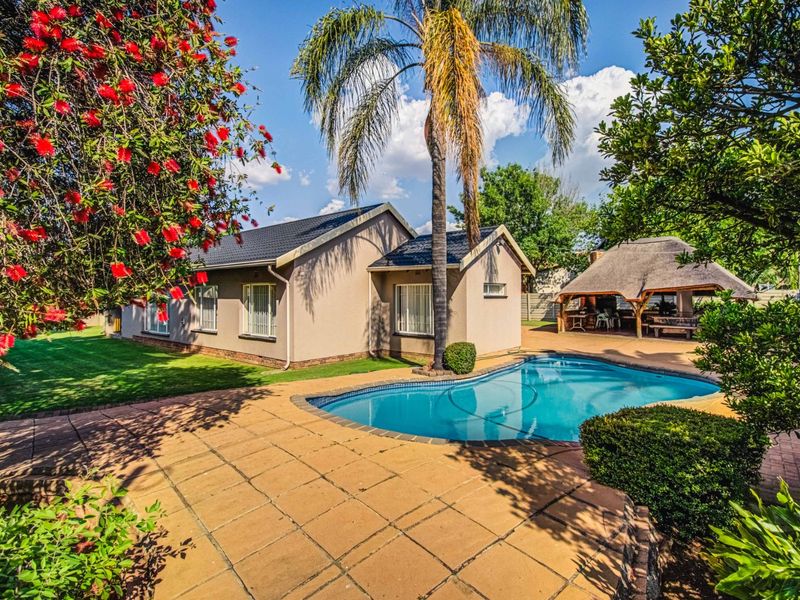 Rear view of the property featuring a well-maintained swimming pool, paved patio area, and a traditional thatched lapa set within a landscaped garden.