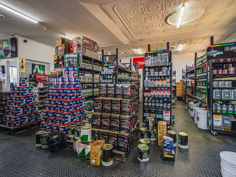 Commercial retail space featuring industrial rubber flooring, pressed metal ceiling panels, and fluorescent lighting, currently utilized for paint and automotive supply storage.