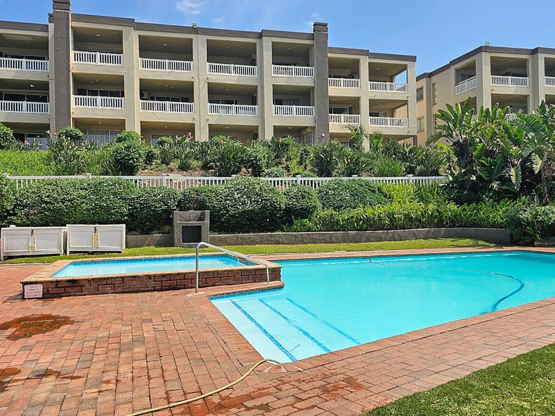 Communal swimming pool area featuring brick paving, a built-in braai, and landscaped gardens set against the apartment complex facade.