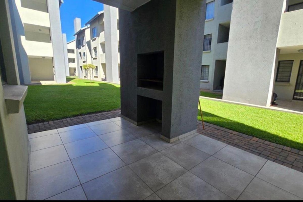 A covered patio area featuring tiled flooring and a built-in braai, overlooking a communal garden space within the apartment complex.