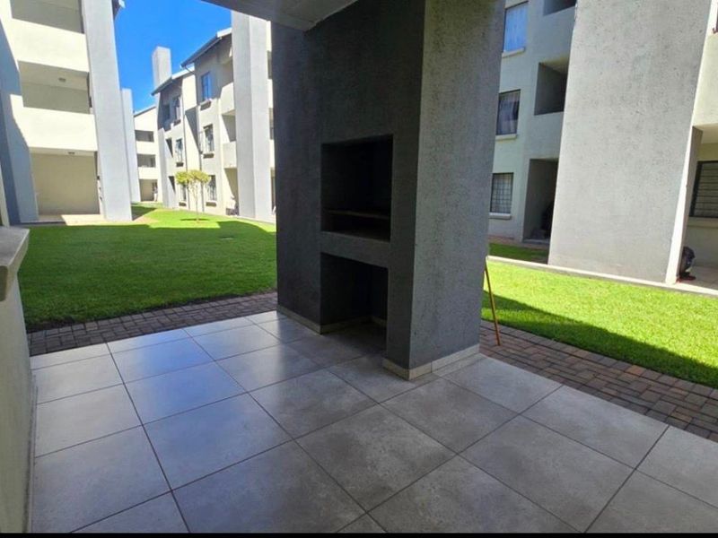 A covered patio area featuring tiled flooring and a built-in braai, overlooking a communal garden space within the apartment complex.