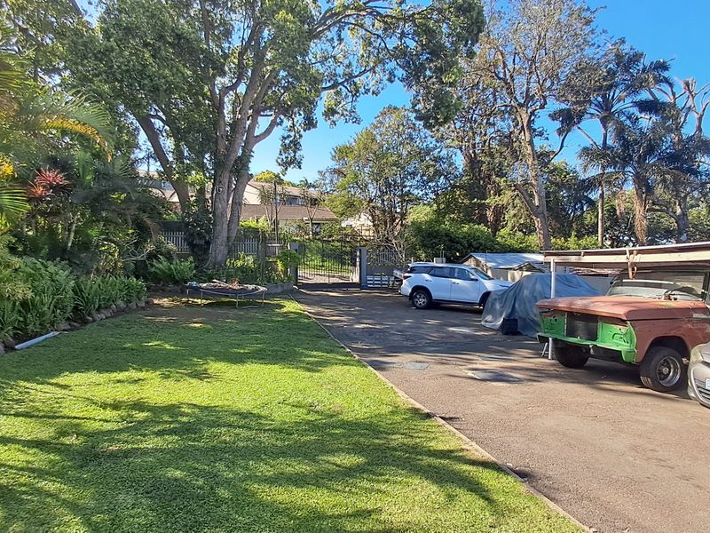 A spacious front exterior featuring a paved driveway, a covered carport area, and a well-established garden with mature trees.