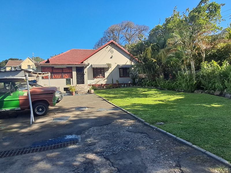Front view of a single-story house featuring a tiled roof, security gate, window awnings, and a large front lawn with a paved driveway.