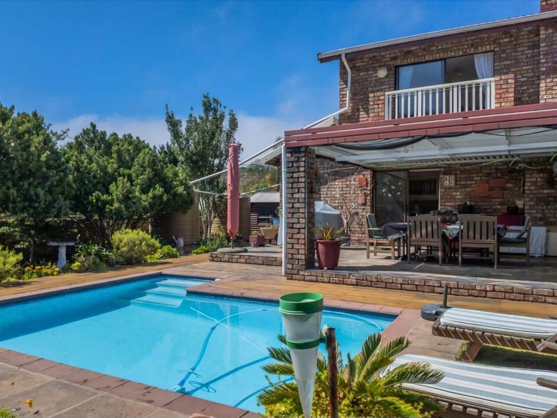 A well-maintained outdoor entertainment area featuring a swimming pool, wooden decking, and a covered patio attached to a brick-faced two-story home.