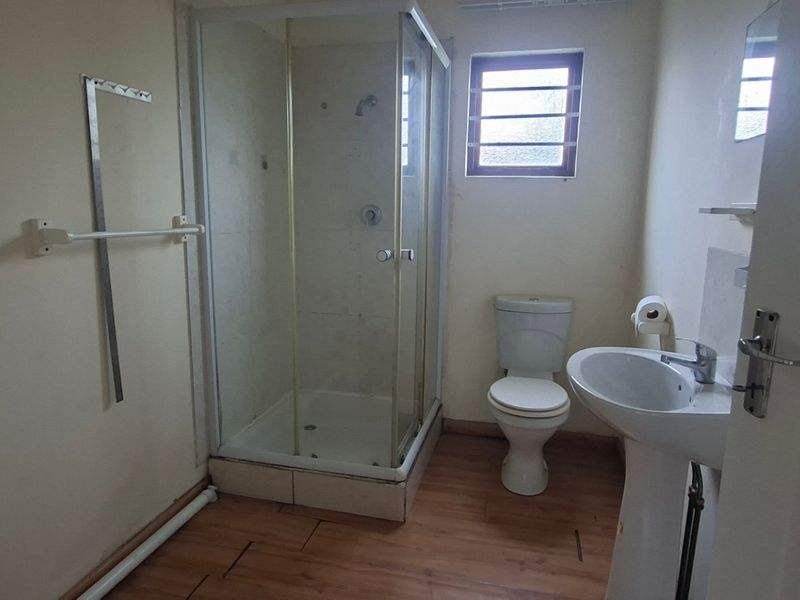 A functional bathroom featuring a shower enclosure, pedestal basin, and toilet, with laminate flooring and a window with security bars.
