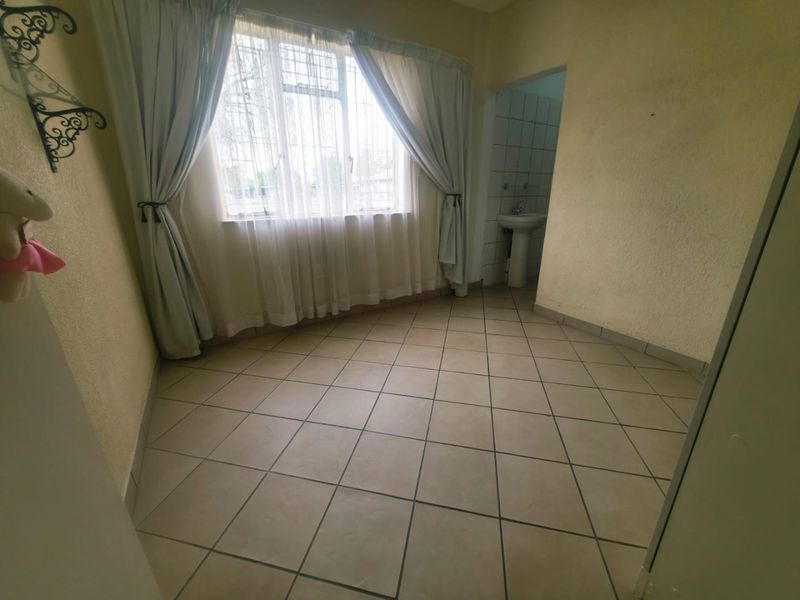 An empty bedroom featuring tiled flooring, a window with security bars, and an adjoining small wash area with a pedestal basin.