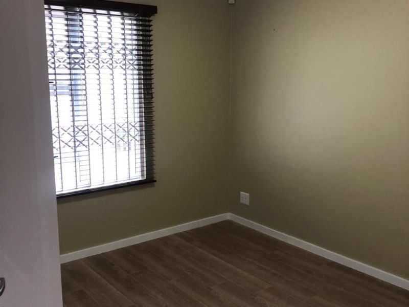 A clean, empty bedroom featuring modern laminate flooring, neutral wall tones, and a window fitted with a security gate and venetian blinds.