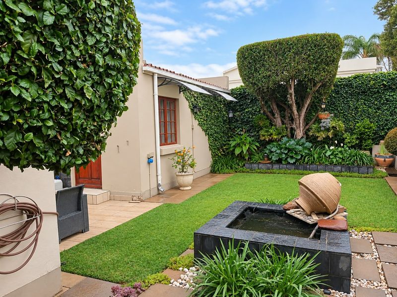 A well-maintained private garden featuring a built-in water feature, manicured lawn, established greenery, and a paved patio area adjacent to the home.