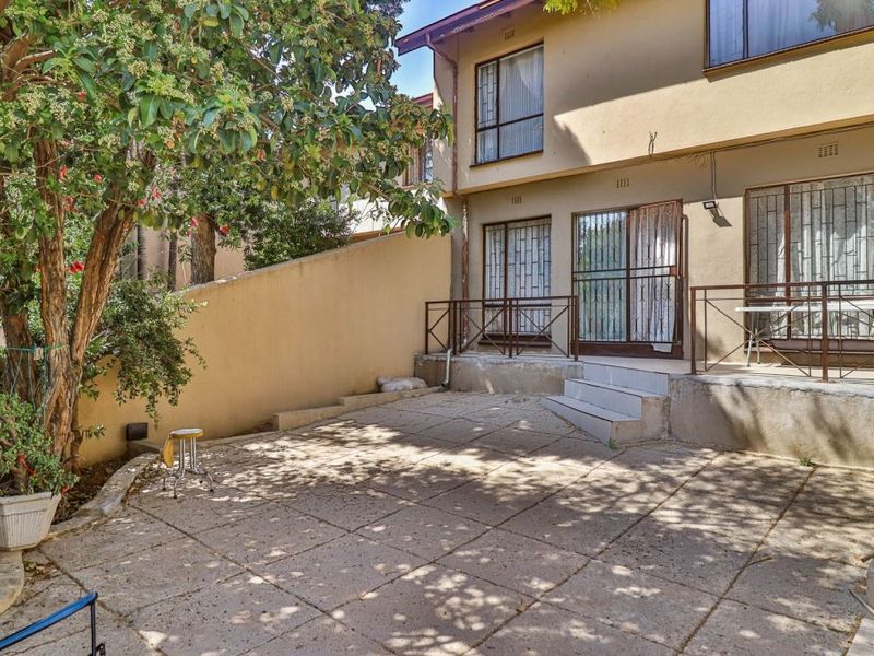 Rear exterior view of the apartment featuring a paved patio area, retaining wall, and metal security gates leading into the living space.