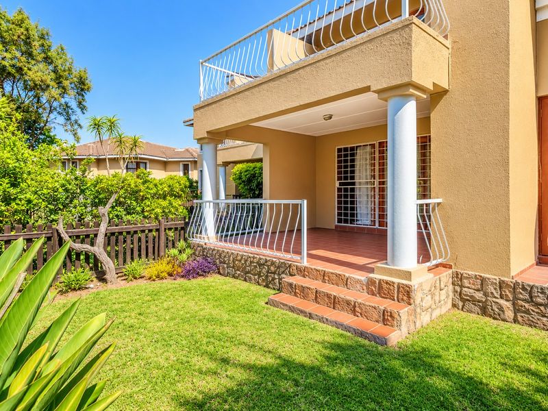 A well-maintained covered patio featuring tiled flooring, stone cladding, and white metal balustrades, overlooking a private garden area.