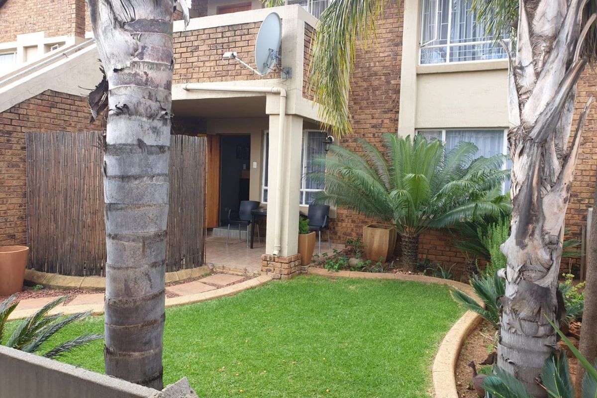 Rear view of a townhouse featuring a brick facade, a covered patio area, and a well-maintained private garden with mature palm trees.
