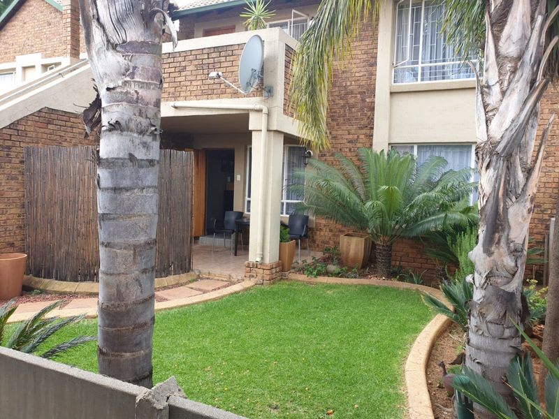 Rear view of a townhouse featuring a brick facade, a covered patio area, and a well-maintained private garden with mature palm trees.