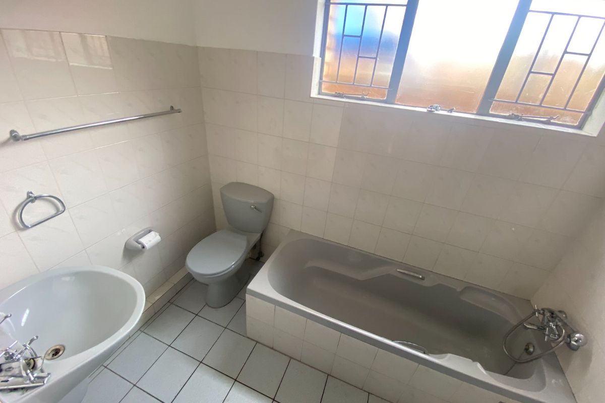A functional bathroom featuring a pedestal basin, toilet, and built-in bathtub with tiled finishes and a window with security bars.