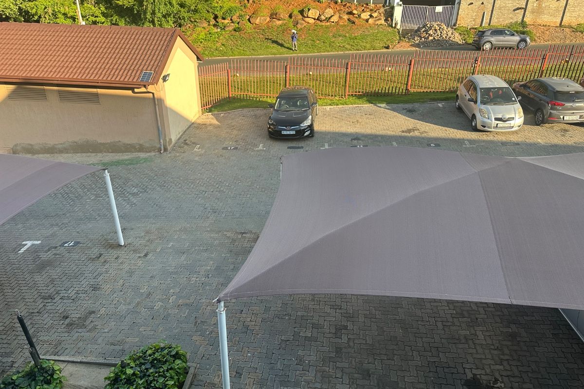 An elevated view of a paved parking area featuring shade cloth carports and perimeter fencing within the apartment complex grounds.