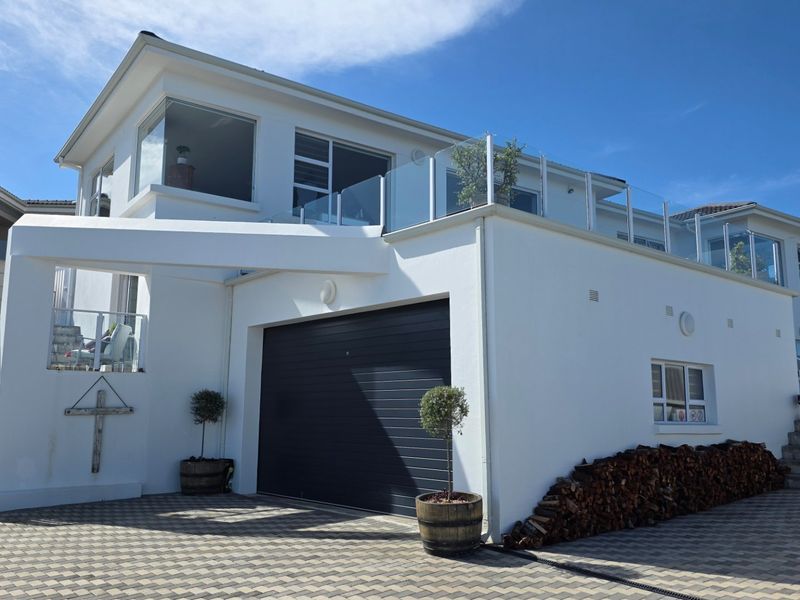 Modern double-story residential facade featuring a clean white finish, automated garage door, paved driveway, and an elevated glass-railed balcony.