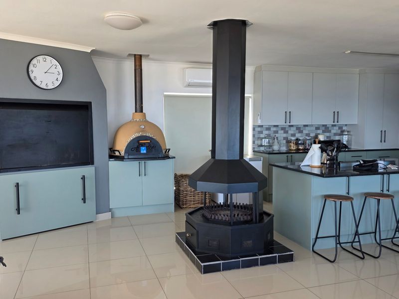 Spacious open-plan living area featuring a modern kitchen with island, built-in braai, pizza oven, and a central fireplace, finished with sleek tiled flooring.