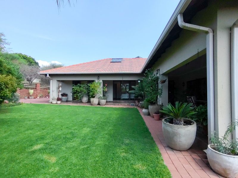 A well-maintained rear exterior view featuring a spacious lawn, brick paved pathways, and a covered patio area with terracotta roof tiles.
