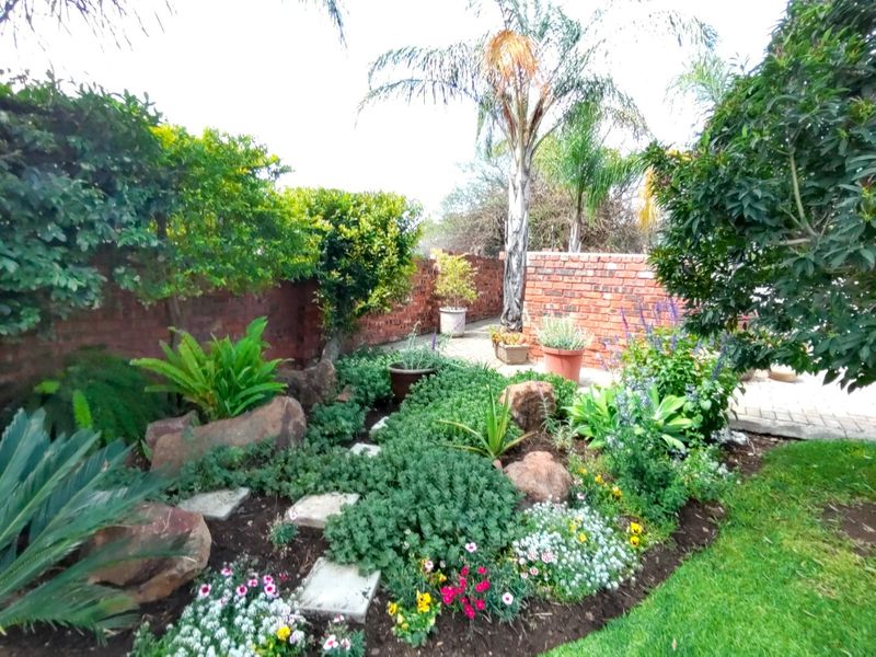 A well-maintained garden featuring landscaped flower beds, mature trees, a paved walkway, and a classic brick boundary wall.