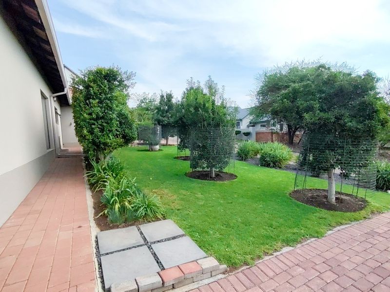 A well-maintained rear garden featuring a manicured lawn, mature trees, and a paved brick walkway along the side of the house.