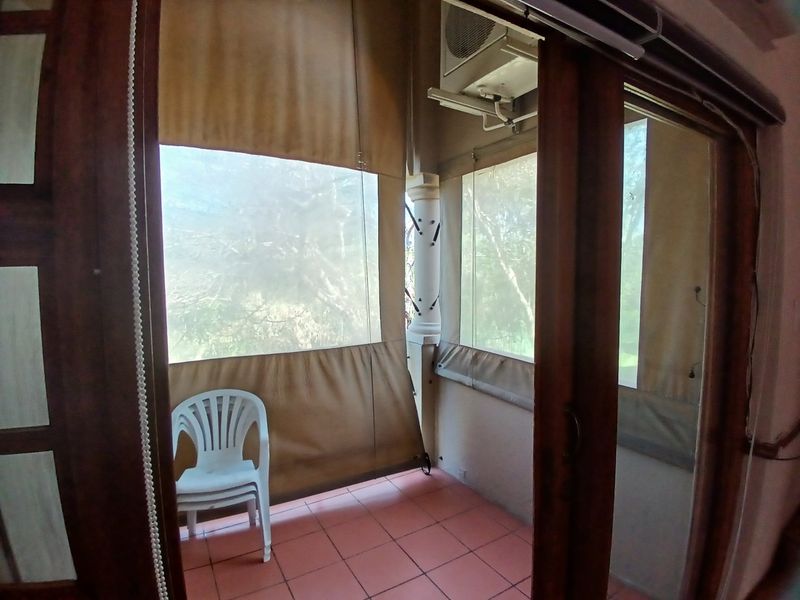 A covered balcony featuring tiled flooring, outdoor roller blinds for privacy, and a wall-mounted air conditioning unit, accessible via sliding glass doors.