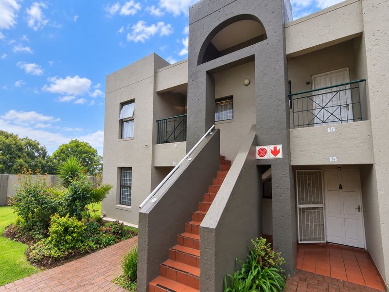 Exterior view of a multi-story apartment complex featuring a central staircase, private balconies, and a well-maintained paved entrance area with landscaped garden beds.
