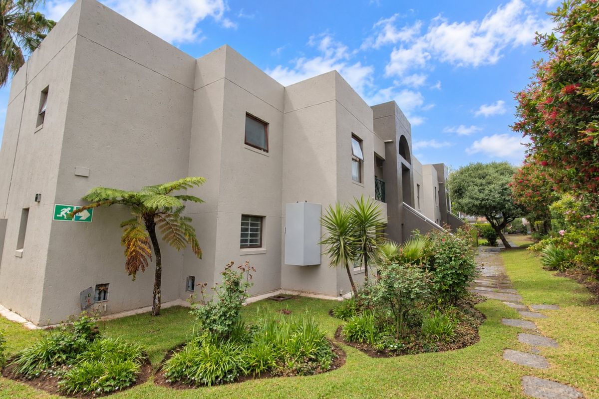A view of the apartment building's rear exterior, featuring a well-maintained stucco facade, landscaped garden beds, and a stone pathway.