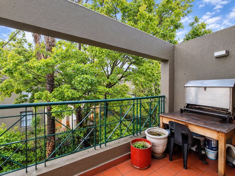 A private balcony featuring tiled flooring, a metal balustrade, and a structural concrete beam, offering a pleasant outlook over mature trees.