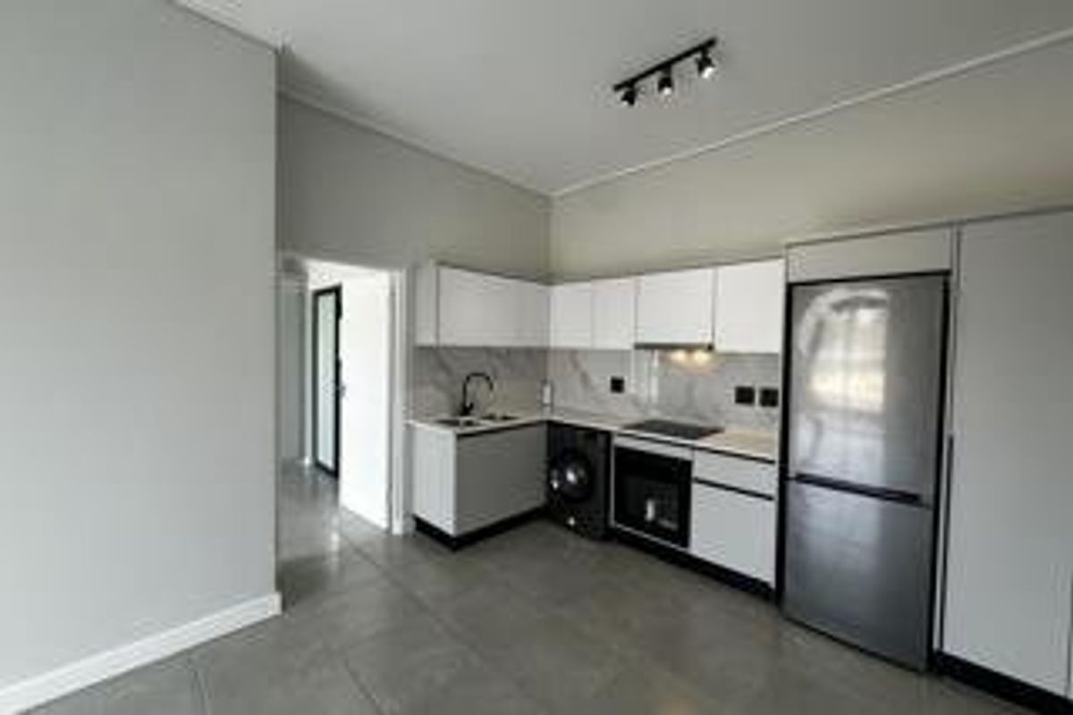 Modern, minimalist kitchen featuring sleek white cabinetry, stone countertops, integrated appliances, and contemporary track lighting in an open-plan layout.