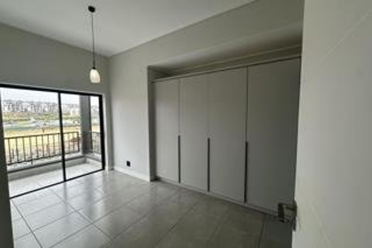 A bright, modern bedroom featuring neutral tiled flooring, extensive built-in cupboards, and a sliding glass door providing access to a private balcony.