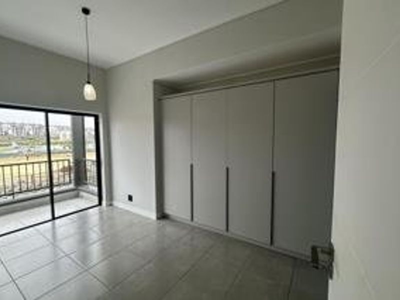 A bright, modern bedroom featuring neutral tiled flooring, extensive built-in cupboards, and a sliding glass door providing access to a private balcony.