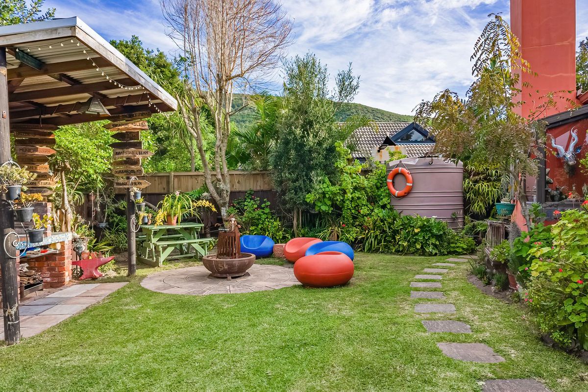 A landscaped rear garden featuring a covered patio with a built-in brick braai, stone pathways, and a water tank.