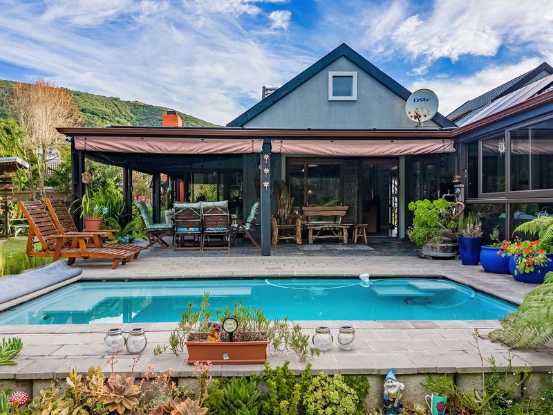 A well-maintained outdoor pool area featuring a paved deck, a covered patio with a retractable awning, and established garden landscaping.