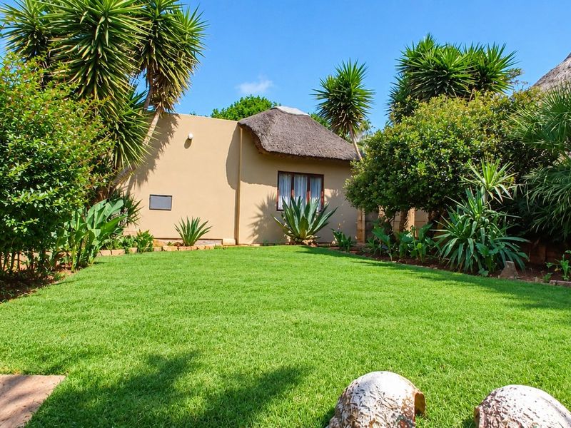 A well-maintained garden featuring a lush green lawn, mature tropical landscaping, and a charming structure with a traditional thatched roof.